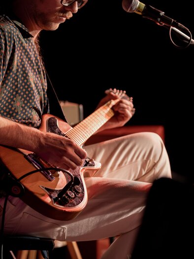 Ein Musiker spielt Gitarre auf der Bühne. Der Fokus liegt auf seinen Händen und dem Instrument. | © Achromatic Photography
