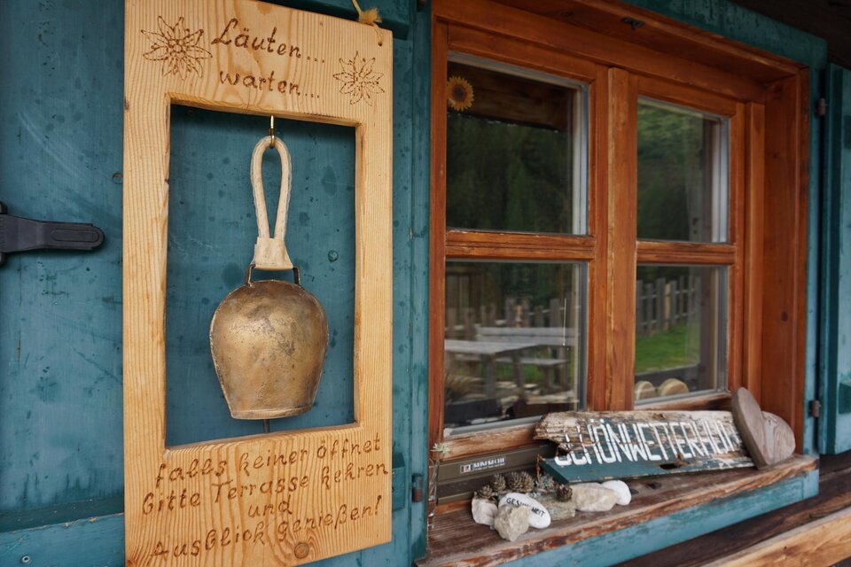 Eine kleine Holztafel mit einer Handglocke hängt an der Wand. Neben dem Fenster sind ebenfalls Hinweise und Steine zu sehen. | © Naturpark Sölktäler