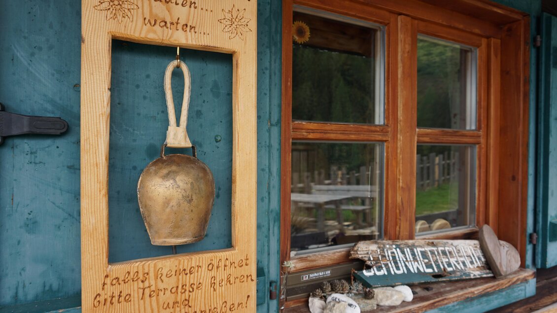 Eine kleine Holztafel mit einer Handglocke hängt an der Wand. Neben dem Fenster sind ebenfalls Hinweise und Steine zu sehen. | © Naturpark Sölktäler