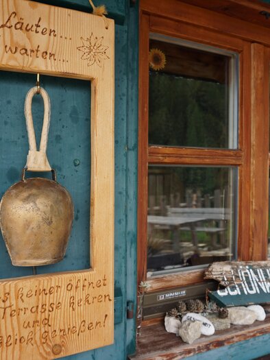 Eine kleine Holztafel mit einer Handglocke hängt an der Wand. Neben dem Fenster sind ebenfalls Hinweise und Steine zu sehen. | © Naturpark Sölktäler