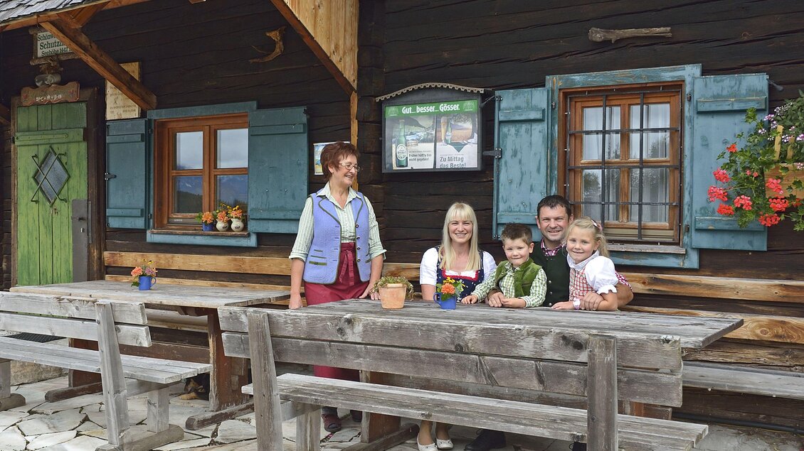 Eine Familie steht vor einem rustikalen Holzhaus mit blauen Fensterläden. Der Tisch ist mit Topfpflanzen dekoriert und zeigt eine einladende Atmosphäre. | © Herfried Marek