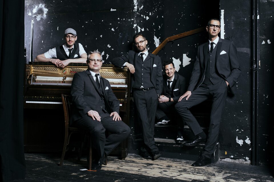 Eine Band aus fünf Männern in eleganter Kleidung steht vor einem alten Klavier. Der Hintergrund ist dunkel und hat einen rauen, unvollkommenen Look. | © Oststeiermark Tourismus