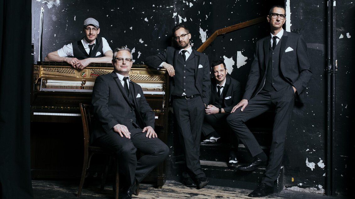 Eine Band aus fünf Männern in eleganter Kleidung steht vor einem alten Klavier. Der Hintergrund ist dunkel und hat einen rauen, unvollkommenen Look. | © Oststeiermark Tourismus