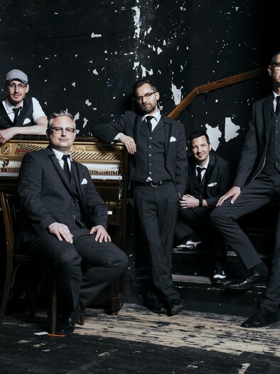 Eine Band aus fünf Männern in eleganter Kleidung steht vor einem alten Klavier. Der Hintergrund ist dunkel und hat einen rauen, unvollkommenen Look. | © Oststeiermark Tourismus