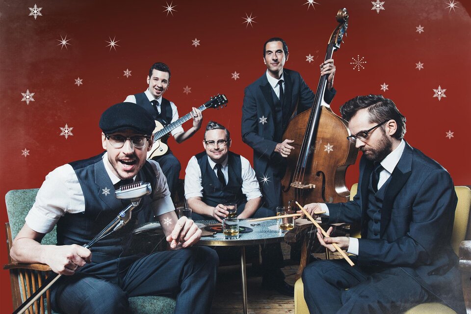 Eine Band in eleganter Kleidung spielt Musik in einem festlichen Raum mit rotem Hintergrund und Schneeflocken. Sie sitzen um einen Tisch mit Getränken und Instrumenten.