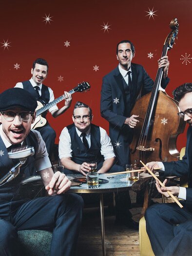 A band in elegant clothing is playing music in a festive room with a red background and snowflakes. They are sitting around a table with drinks and instruments.