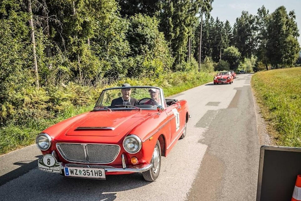 Zwei rote Oldtimer fahren auf einer ruhigen Landstraße. Umgeben von grünen Bäumen und sonnigem Wetter. | © Ewald Schneeberger