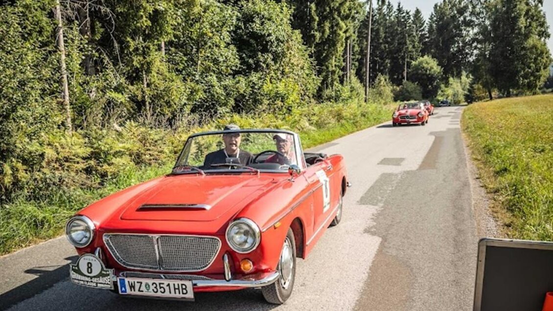 Zwei rote Oldtimer fahren auf einer ruhigen Landstraße. Umgeben von grünen Bäumen und sonnigem Wetter. | © Ewald Schneeberger