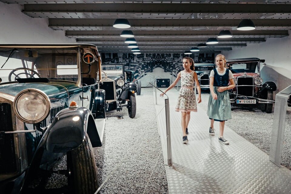 Zwei Mädchen gehen durch ein Automuseum und betrachten historische Fahrzeuge. Die Umgebung ist modern gestaltet mit einer hellen und einladenden Atmosphäre.