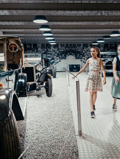 Zwei Mädchen gehen durch ein Automuseum und betrachten historische Fahrzeuge. Die Umgebung ist modern gestaltet mit einer hellen und einladenden Atmosphäre.
