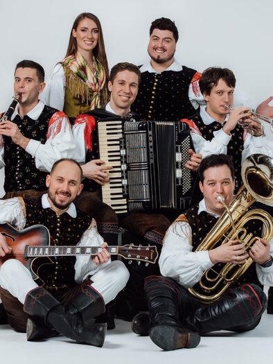 Eine Gruppe von sieben Musikern in traditioneller Tracht. Sie spielen verschiedene Instrumente und posieren fröhlich vor einem hellen Hintergrund. | © avsenik