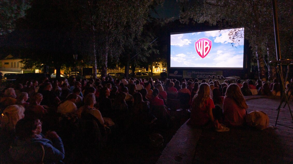 Eine Menschenmenge sitzt im Freien und schaut einen Film auf einer großen Leinwand. Das Logo von Warner Bros ist im Hintergrund zu sehen. | © Christoph Graif