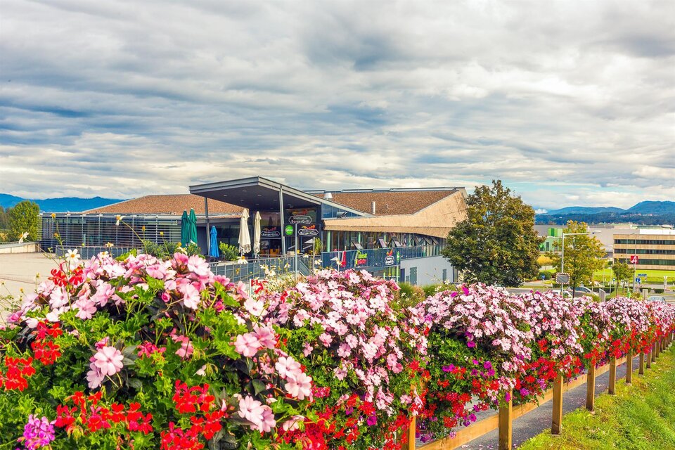 Ein modernes Gebäude umgeben von bunten Blumen. Der Himmel ist bewölkt und es deutet sich ein schöner Tag an. | © MG Lannach