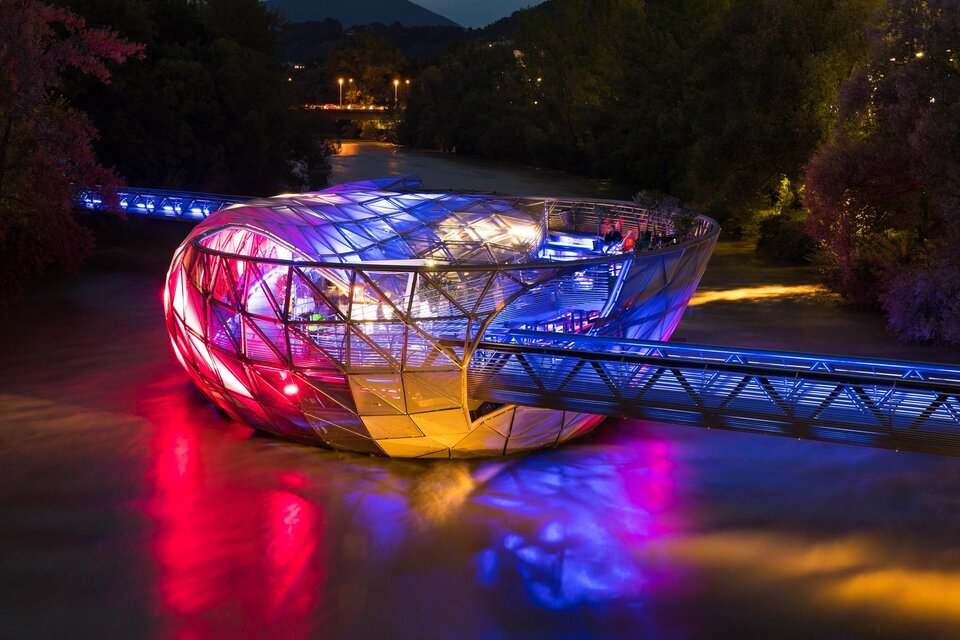 Eine moderne, beleuchtete Brücke mit einer besonderen, futuristischen Form über einem Fluss. Die Farben und Lichter reflektieren sich im Wasser. | © Graz Tourismus - Harry Schiffer