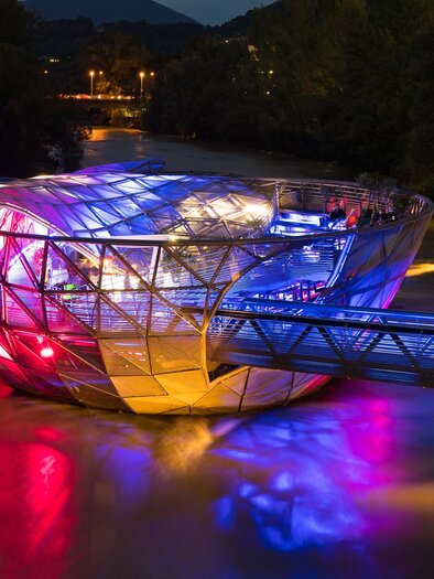 Eine moderne, beleuchtete Brücke mit einer besonderen, futuristischen Form über einem Fluss. Die Farben und Lichter reflektieren sich im Wasser. | © Graz Tourismus - Harry Schiffer