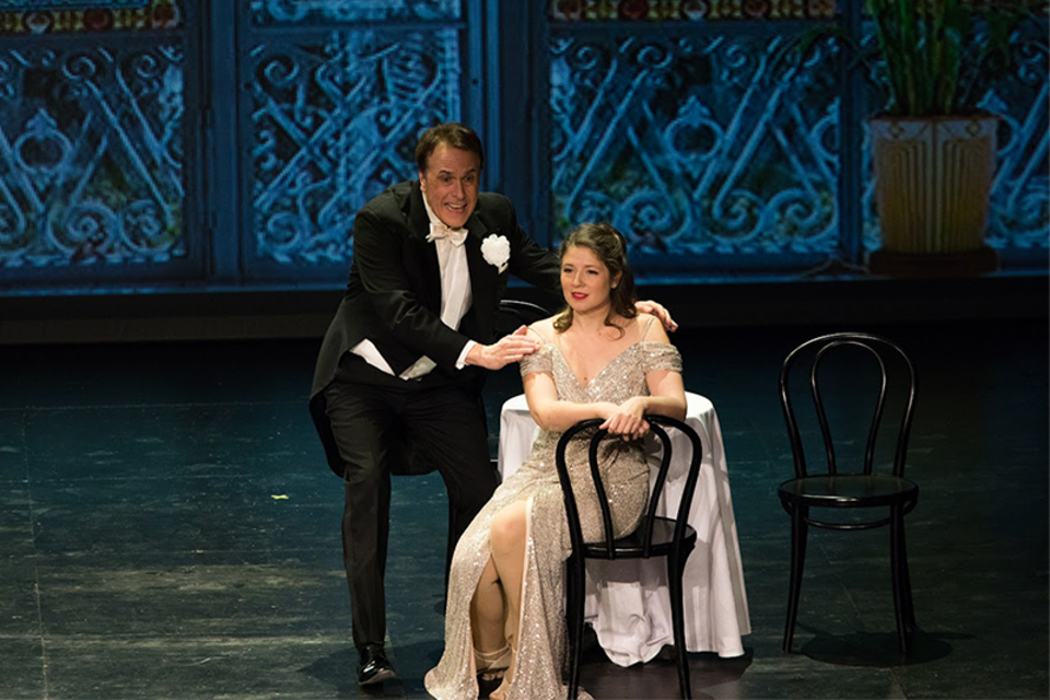 Ein schauspielerisches Paar steht auf der Bühne, der Mann in eleganter Abendkleidung und die Frau in einem glitzernden Kleid. Sie sitzen an einem Tisch mit zwei Stühlen, während im Hintergrund eine dekorative Kulisse zu sehen ist. | © Schutte