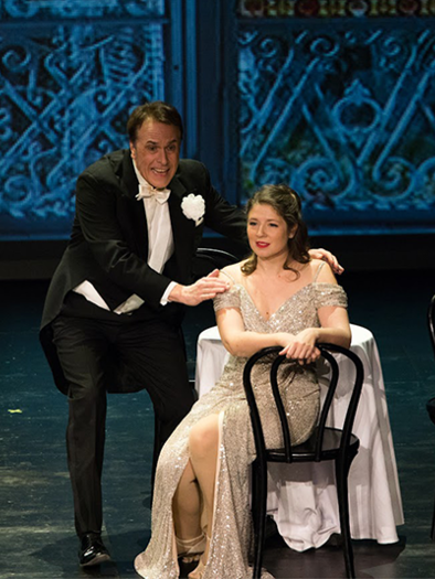 An acting couple stands on stage, the man in elegant evening wear and the woman in a sparkling dress. They are sitting at a table with two chairs, while a decorative backdrop is visible in the background. | © Schutte