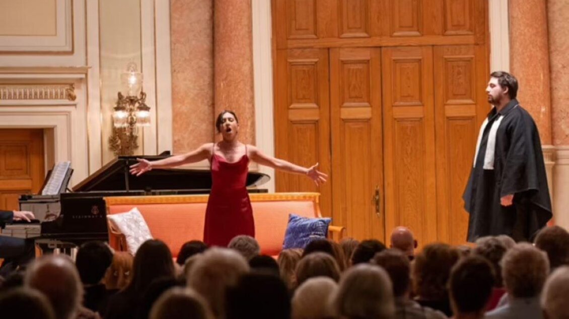 Eine Sängerin in einem roten Kleid tritt zusammen mit einem Sänger in einem Anzug auf. Der Auftritt findet in einem eleganten Saal mit einem Klavier statt, während das Publikum aufmerksam zuhört. | © Gerald Hirl