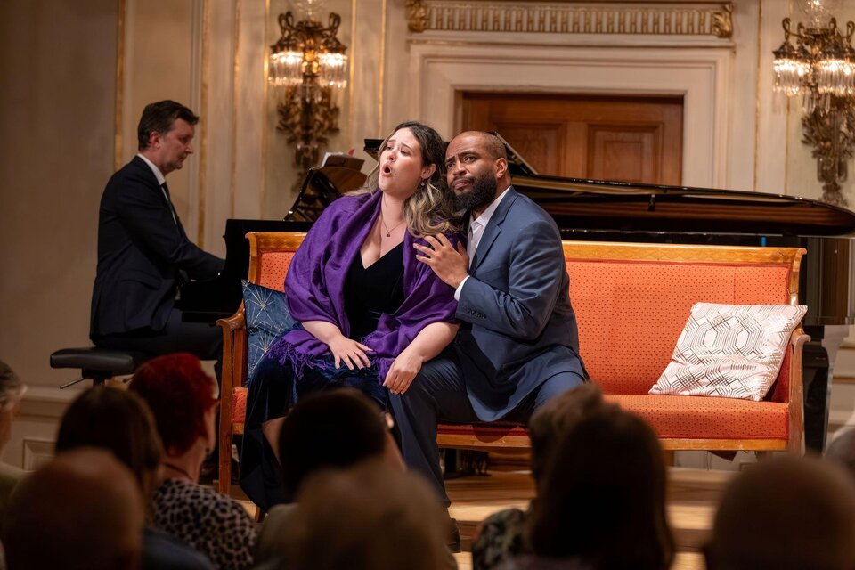 Eine Aufführung mit zwei Sängern, die auf einer orangefarbenen Couch sitzen. Im Hintergrund spielt ein Pianist. | © AIMS Gerald Hirl