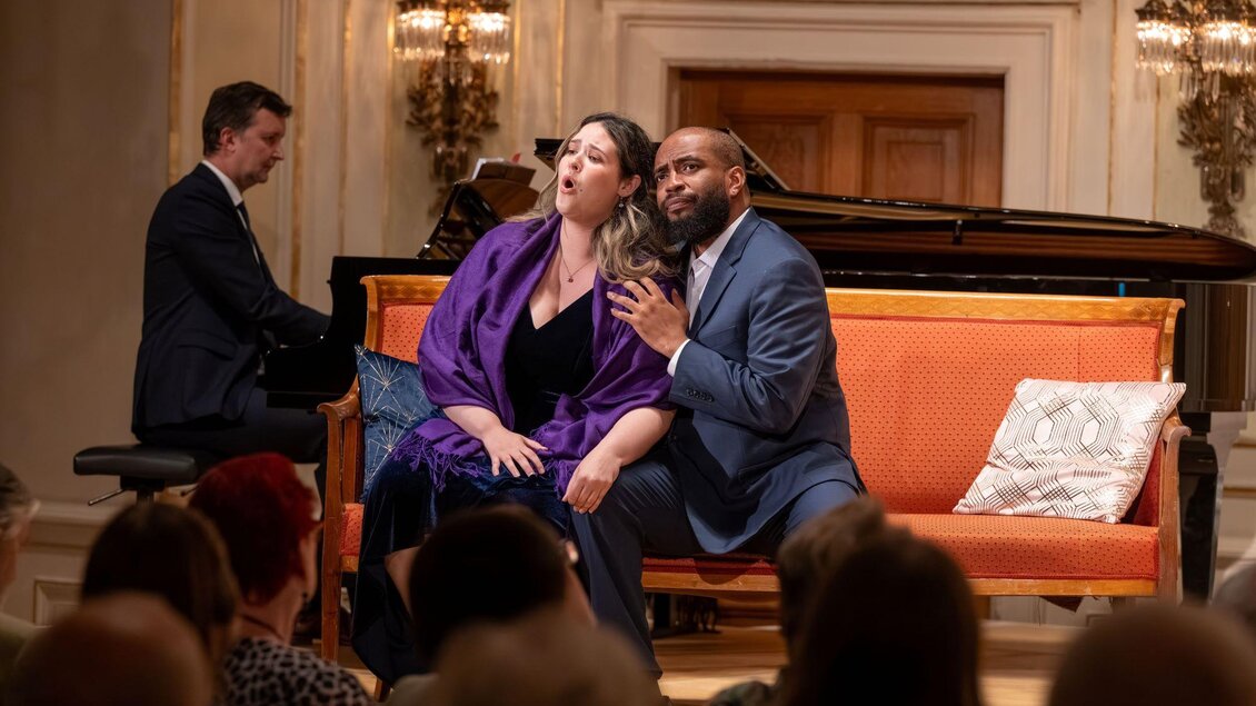 Eine Aufführung mit zwei Sängern, die auf einer orangefarbenen Couch sitzen. Im Hintergrund spielt ein Pianist. | © AIMS Gerald Hirl
