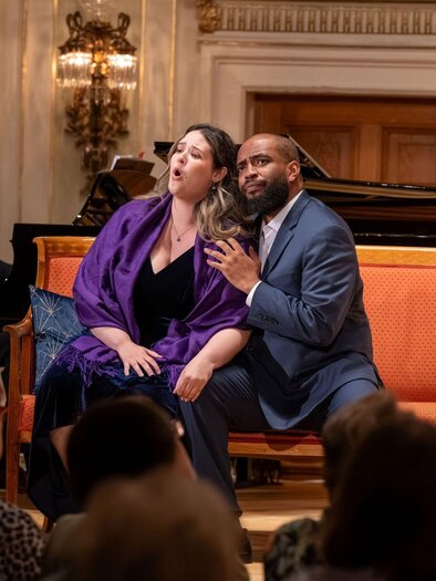 Eine Aufführung mit zwei Sängern, die auf einer orangefarbenen Couch sitzen. Im Hintergrund spielt ein Pianist. | © AIMS Gerald Hirl