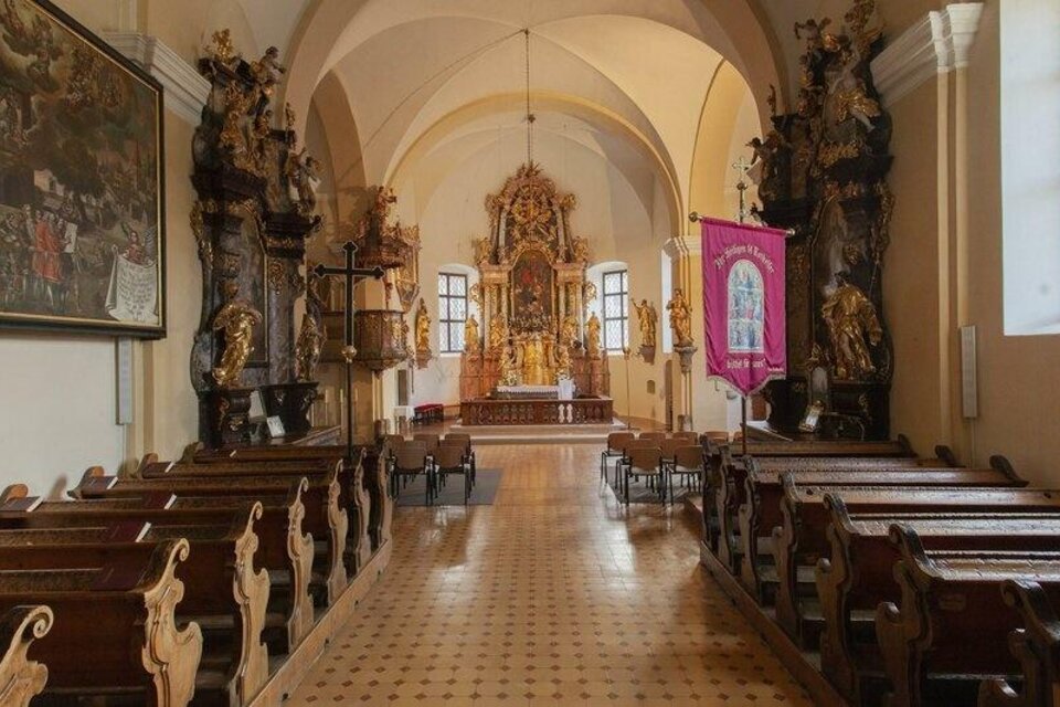 Eine stille Kirche mit Holzbankreihen und einem prächtigen Altar im Hintergrund. Die Wände sind reich verziert und es gibt große Fenster, die Licht hineinlassen. | © Pollhammer