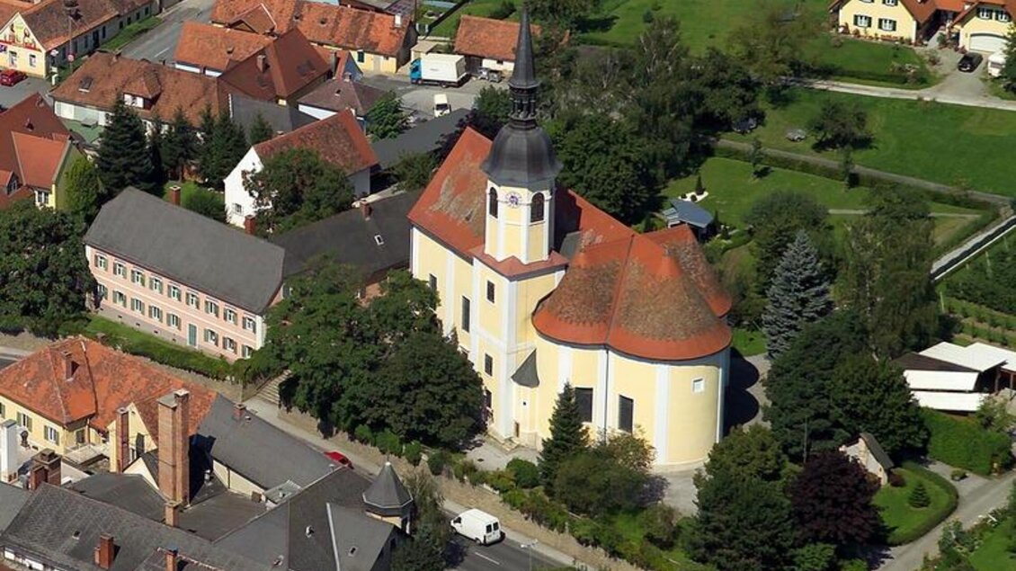 Eine malerische Kirche mit einem hohen Turm, umgeben von grünen Bäumen. Rundum sind typische Häuser und eine ruhige Landschaft zu sehen. | © Pfarre Kaindorf/Scheiblhofer Rainer