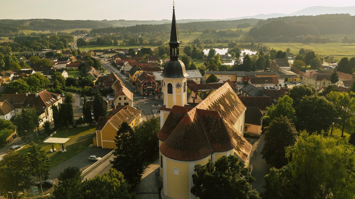 Eine malerische Stadtansicht mit einer Kirche und einem Glockenturm. Die Umgebung ist grün und hügelig, umgeben von Bäumen und einem ruhigen Fluss. | © Oststeiermark Tourismus