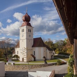 Eine malerische Kirche mit einem markanten Turm und roten Dächern. Umgeben von einer grünen Landschaft und einem klaren Himmel.