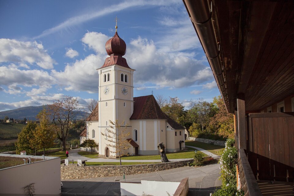 Eine malerische Kirche mit einem markanten Turm und roten Dächern. Umgeben von einer grünen Landschaft und einem klaren Himmel.