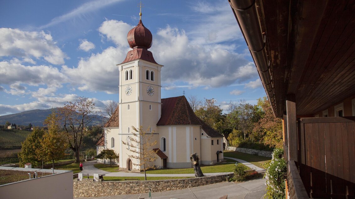 Eine malerische Kirche mit einem markanten Turm und roten Dächern. Umgeben von einer grünen Landschaft und einem klaren Himmel.