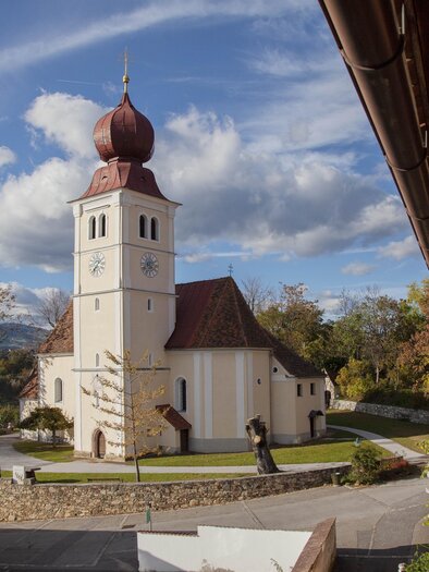 Eine malerische Kirche mit einem markanten Turm und roten Dächern. Umgeben von einer grünen Landschaft und einem klaren Himmel.