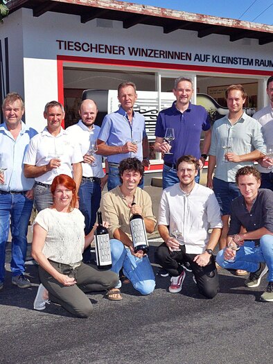 A group of men and women are standing in front of a building, holding bottles in their hands. They are smiling and appear cheerful as they pose together. | © TAU-Winzer Tieschen