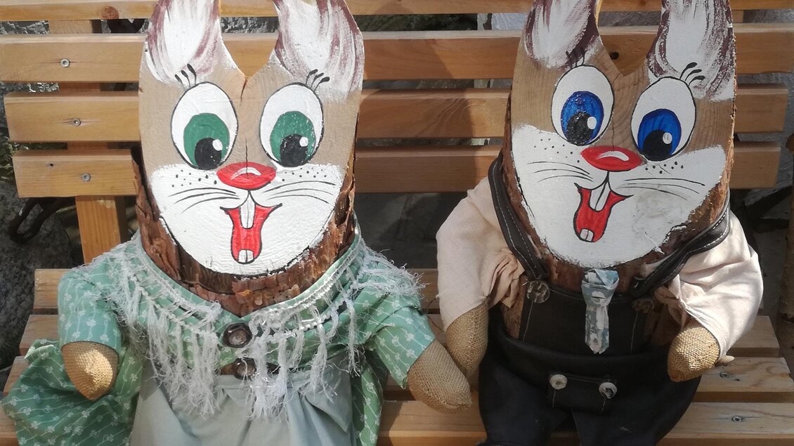 Zwei dekorative Figuren in Form von Hasen sitzen auf einer Holzbank. Die linke Figur trägt ein grünes Kleid, während die rechte Figur eine schwarze Weste anhat. | © Tourismusverein Fischbach