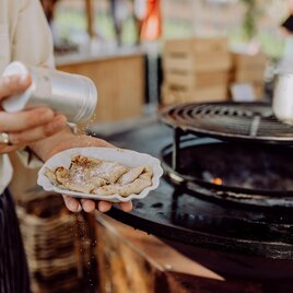 A cook is seasoning a portion of food on a plate. In the background, a grill and a cozy atmosphere can be seen. | © Karin Bergmann