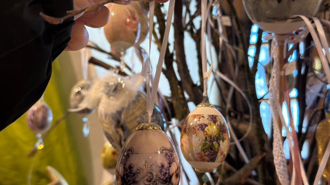 Eine Hand hält ein dekoratives Osterei, das an einem kunstvoll geschmückten Ast hängt. Verschiedene bunte Eier verzieren den Zweig und schaffen eine festliche Atmosphäre. | © Schloss Kornberg