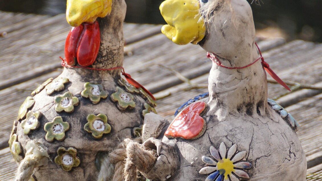 Zwei bunte, dekorative Hühner aus Ton stehen auf einem Holzbrett. Sie haben lustige Gesichter und sind mit Blumenmuster verziert. | © Kunsthandwerk Schloss Kornberg, Leonhard Bardeau