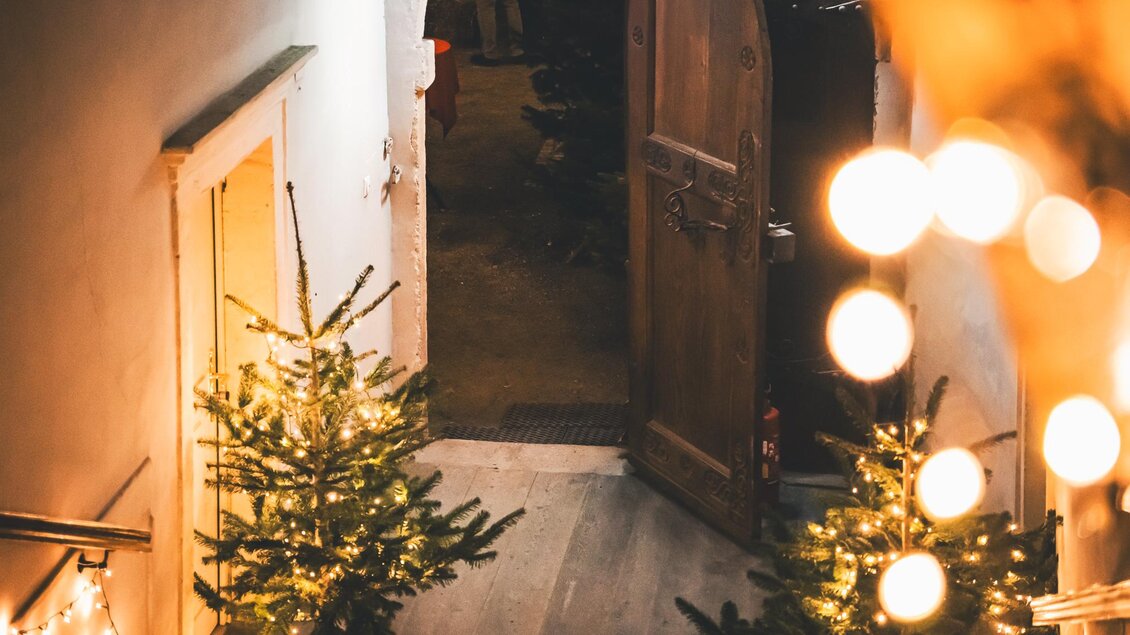 Eine festlich dekorierte Treppe mit leuchtenden Weihnachtsbäumen. Im Hintergrund steht eine geöffnete Tür, die eine warme und einladende Atmosphäre schafft. | © Maria Ebner