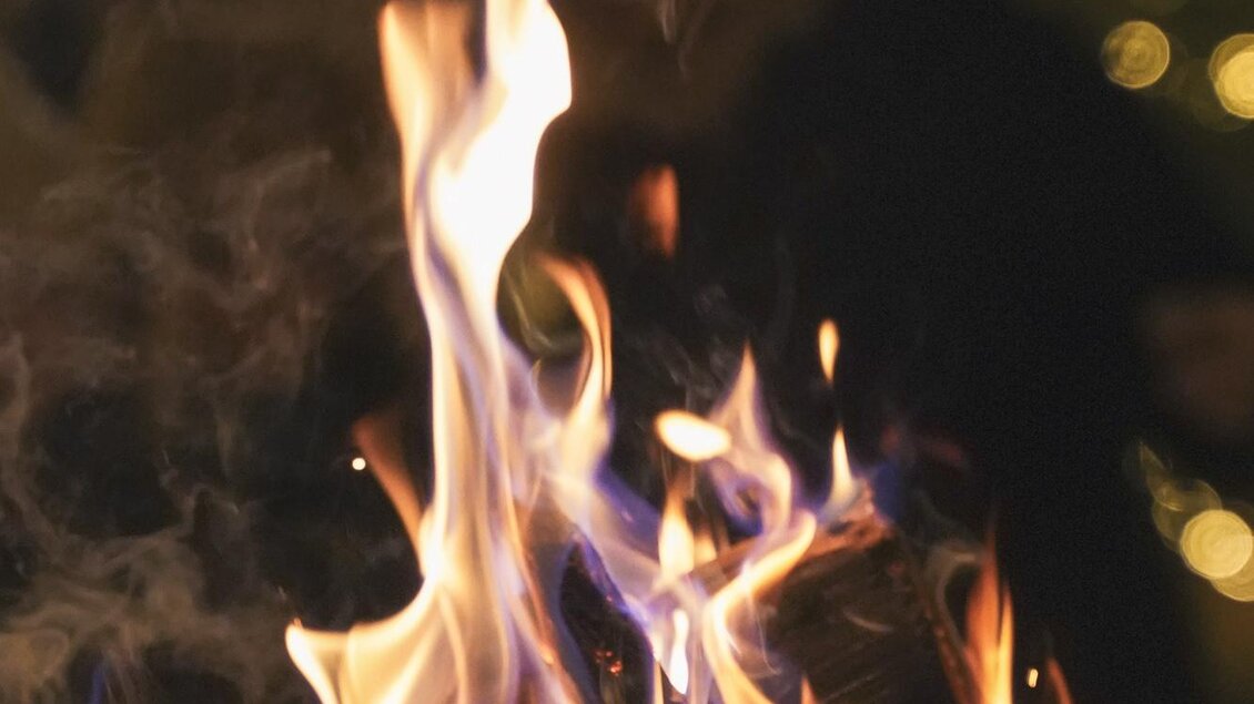 Ein lebhaftes Feuer mit bunten Flammen brennt in einem rustikalen Lager. Im Hintergrund sind unscharfe Lichter und Silhouetten zu sehen. | © Maria Ebner