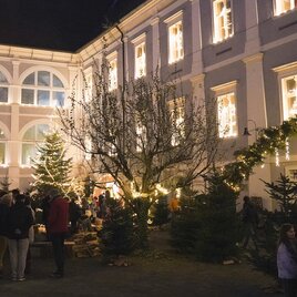 Ein festlicher Weihnachtsmarkt in einem Innenhof mit Lichtern und Tannenbäumen. Menschen stehen zusammen und genießen die Atmosphäre. | © Maria Ebner