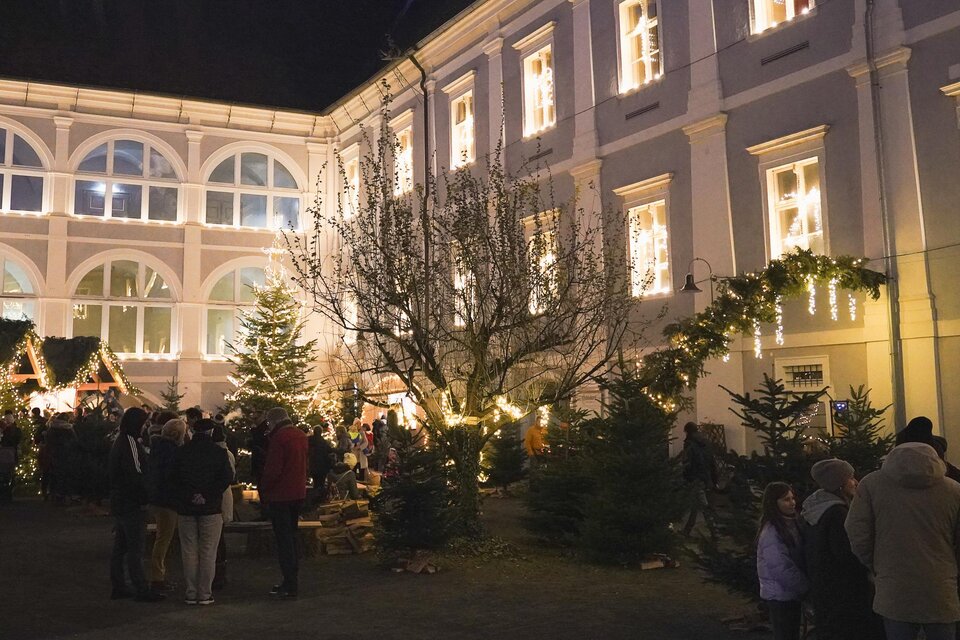 Ein festlicher Weihnachtsmarkt in einem Innenhof mit Lichtern und Tannenbäumen. Menschen stehen zusammen und genießen die Atmosphäre. | © Maria Ebner