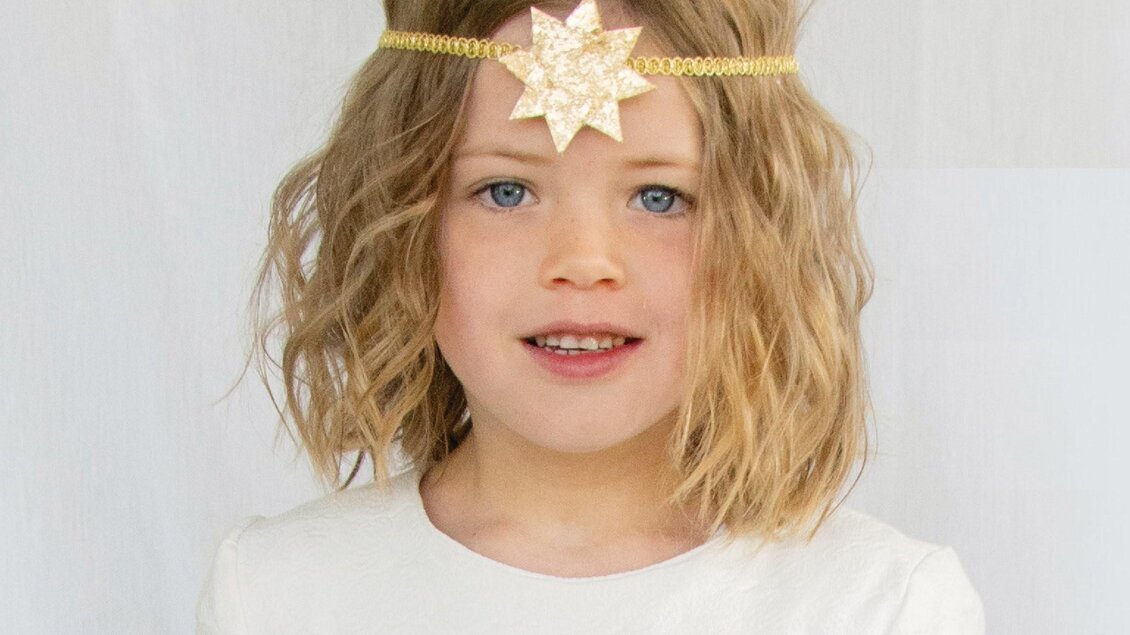 Ein kleines Mädchen mit lockigem Haar trägt ein glitzerndes Stirnband mit einem Stern. Sie lächelt und trägt ein einfaches, helles Kleid. | © Ebner Maria
