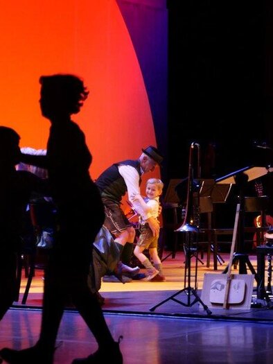 Eine lebhafte Bühnenaufführung mit Musikern und Tänzern. Im Hintergrund leuchtet eine große orangefarbene Wand. | © -styriarte---ct-