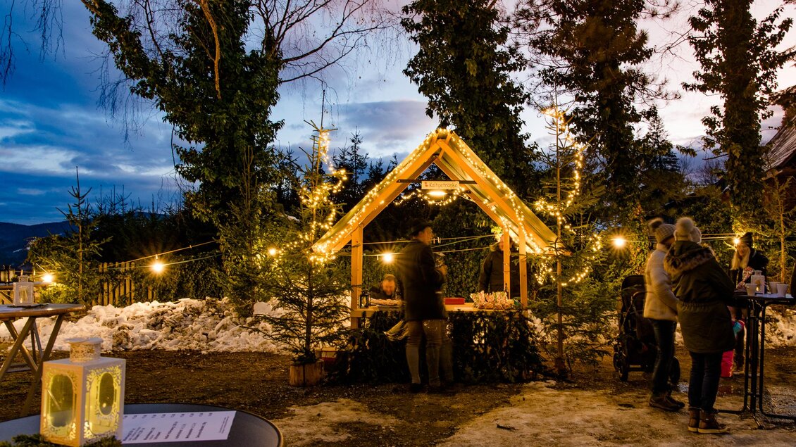 Eine festlich dekorierte Holzhütte steht in einer winterlichen Umgebung. Menschen genießen die Atmosphäre bei Dämmerung und glitzernden Lichtern. | © Region Graz-Mias Photoart