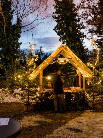 A festively decorated wooden cabin stands in a wintery setting. People enjoy the atmosphere at dusk with sparkling lights. | © Region Graz-Mias Photoart