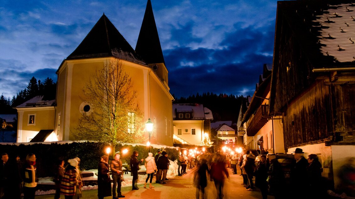 Eine festliche Nachtansicht eines Dorfes mit einer Kirche und vielen Leuten, die Fackeln halten. Der Himmel ist dunkelblau und die Atmosphäre ist warm und einladend. | © Region Graz-Mias Photoart