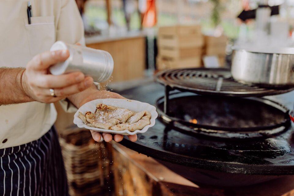 Ein Koch würzt gerade eine Portion Essen auf einem Teller. Im Hintergrund ist ein Grill und eine gemütliche Atmosphäre zu sehen. | © Karin Bergmann