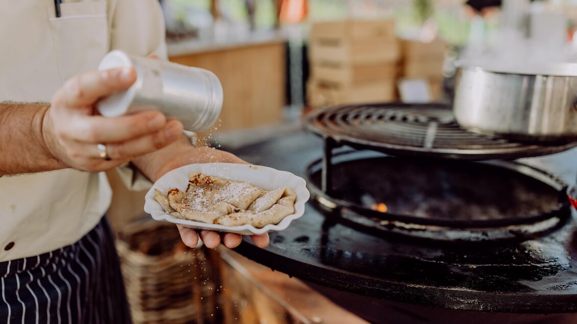 Ein Koch würzt gerade eine Portion Essen auf einem Teller. Im Hintergrund ist ein Grill und eine gemütliche Atmosphäre zu sehen. | © Karin Bergmann