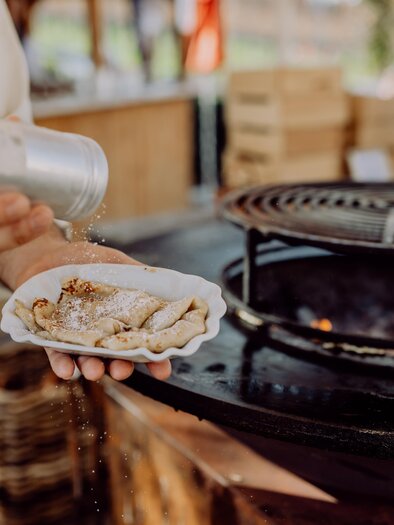 Ein Koch würzt gerade eine Portion Essen auf einem Teller. Im Hintergrund ist ein Grill und eine gemütliche Atmosphäre zu sehen. | © Karin Bergmann