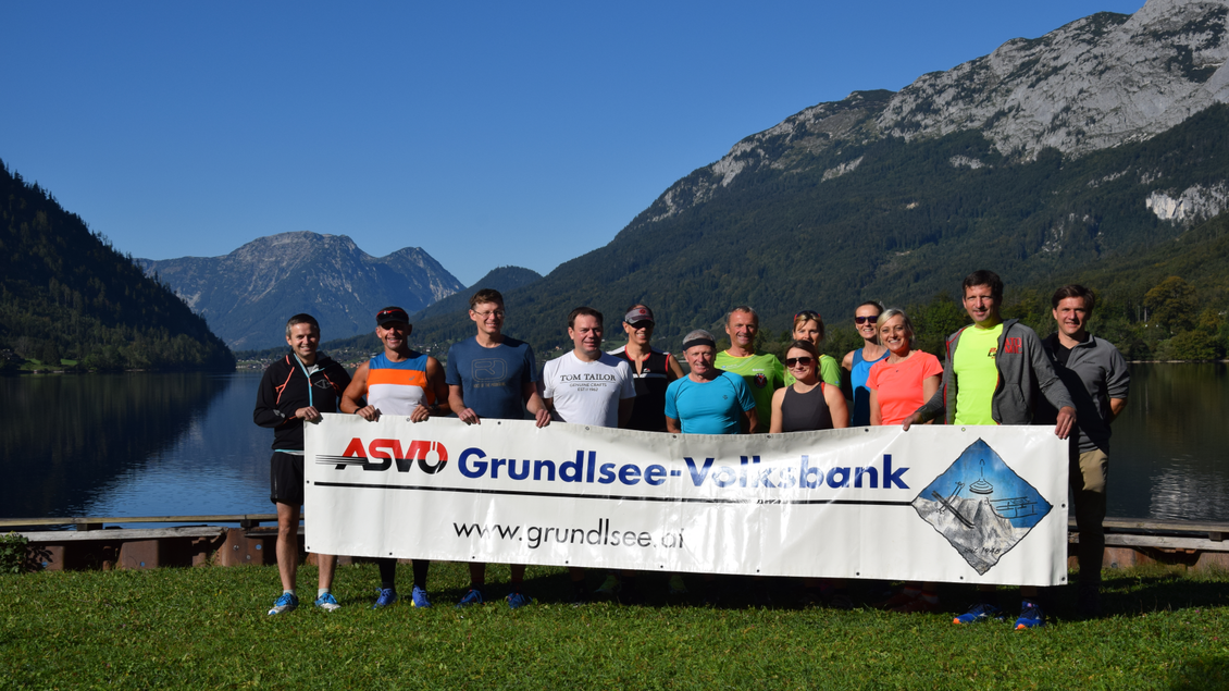 <p>Das Organisationsteam rund um die ASVÖ Grundlsee-Volksbank steht in Gößl mit einem Banner in den Händen und dem Grundlsee im Hintergrund. Die beeindruckende Berglandschaft und der klare Himmel bilden die Kulisse.</p> | © ASVÖ Grundlsee
